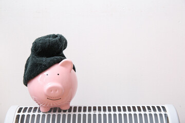 Piggy bank with warm hat on electric convector heater near white wall at home. Heating saving concept