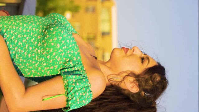 glamorous fancy women model with messy brown hair wearing green cute dress leaning on a fence relaxing her head back with closed eyes and looking straight into camera slow motion vertical cinematic