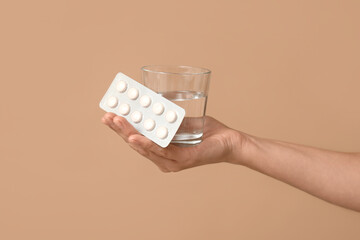 Female hand with pills in blister pack and glass of water on brown background, closeup