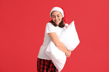 Beautiful young woman in pajamas and Santa hat with pillow on red background