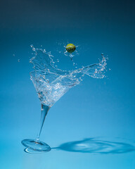 A freshly made Martini cocktail is seen splashing dynamically against a vibrant blue gradient background. An olive garnish hangs above the glass in mid air.