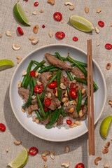 A freshly made dish of green beans, chilli and beef is seen from directly above. Wooden chopsticks sit beside the bowl and wedges of lime rest on the table.