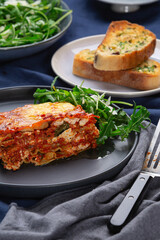 A freshly baked slice of lasagne sits on a plate beside a garden salad. Slices of garlic bread can be seen i the background.