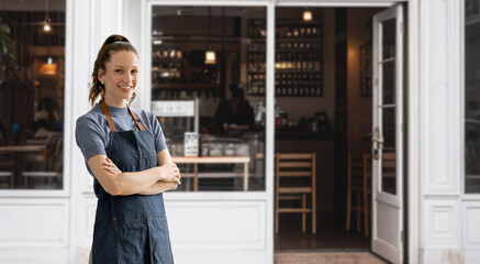 Cheerful young waitress waiting for clients at coffee shop. Successful small business owner in casual wearing jeans apron standing at entrance