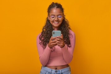 Young optimistic pretty Indian woman teenager holding phone in hands and typing message in college group chat enjoying sharing funny pictures in mobile messenger stands posing in orange studio.