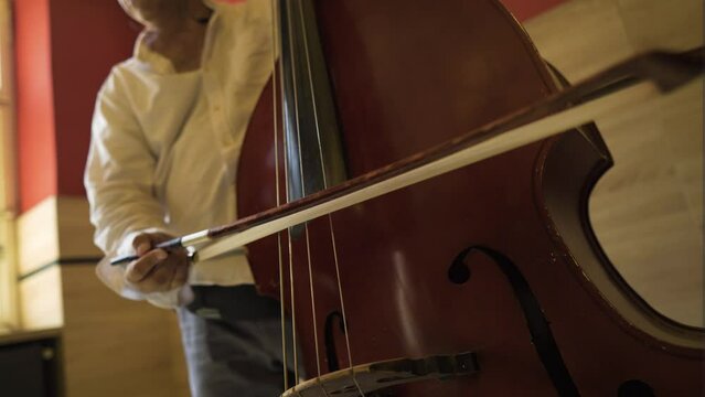 low angle of an olive skin elderly man European playing cello bass with a long stick performing different notes octaves baroque classical royal music gypsy folk style antique instrument white shirt