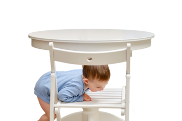 Toddler baby climbs onto a chair at the kitchen white table, isolated on a white background. A child in danger climbs up on the furniture at the risk of falling. Kid aged one year and two months