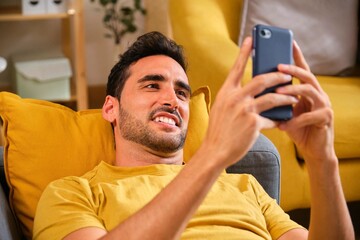 Young smiling Caucasian man using his smartphone laying on the sofa at home.