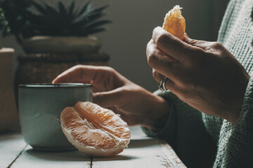 Close up of woman eating orange fruit and drinking herbal tea or coffee at home. healthy food and beverage lifestyle people indoor. Dark mood color green grey. Relaxation and enjoying relax inside - obrazy, fototapety, plakaty