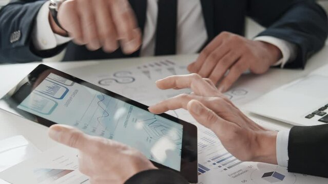 Close up shot of unrecognisable businessmans hands holding tablet with project effectiveness information and discussing it with business associate during office meeting