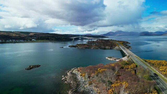 Drone rises above rocky shoreline and yellow fields above islands and Skye bridge in Scotland