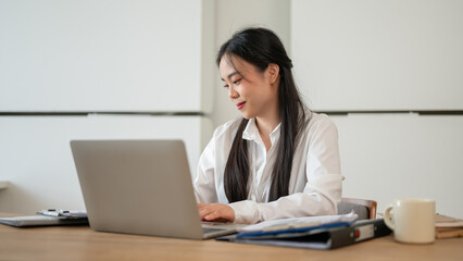 An attractive Asian businesswoman is working on her laptop at her desk in a modern office.