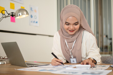 Fototapeta na wymiar A professional Asian-Muslim businesswoman in a hijab is working on financial reports at her desk.