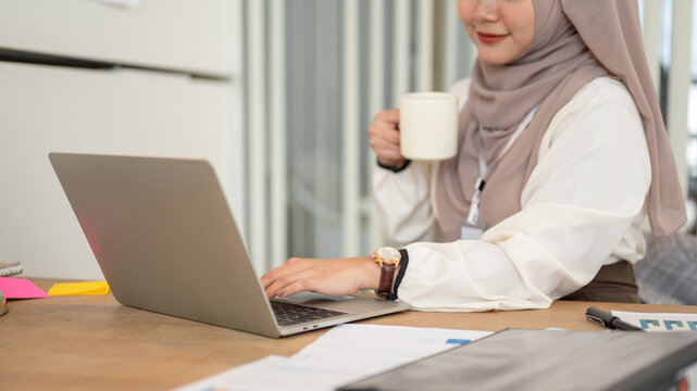 Cropped image of an Asian-Muslim businesswoman is having her coffee while working on her laptop.