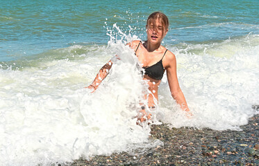 Emotional teenage girl sits in shallow water in foamy stormy sea waves on a sunny warm summer day