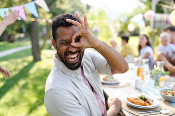 Handsome man making funny faces, sitting at the party table during a summer garden party with...