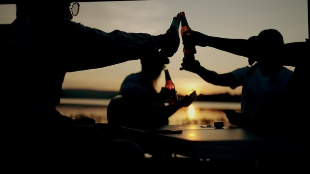 Group of friends playing guitar and drinking beer on silhouette sunset background beautiful nature, mountains and lake,Friendship and celebration concept.