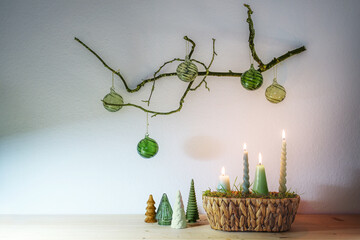 Advent and Christmas decoration with four different candles in a basket and small artificial trees under a hanging bare branch with green glass balls against a light wall, copy space