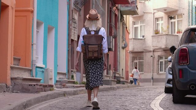 Slow motion beautiful female tourist with backpack in white shirt and straw hat walking along empty street in old tourist part city, enjoying her vacation. Sightseeing in popular tourist destination.