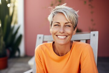 Smiling middle-aged woman sitting on the terrace of her cozy country house. Attractive blonde female with short hairstyle enjoys silence of her beautiful garden. Adult lady with happy smile relaxed. - obrazy, fototapety, plakaty
