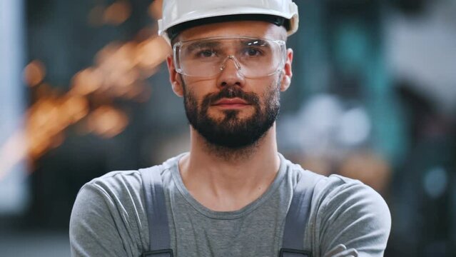 Portrait of a professional heavy industry engineer. Worker wearing protective uniform, goggles and helmet, smiling. In the background is a disoriented large industrial plant where welding sparks fly.
