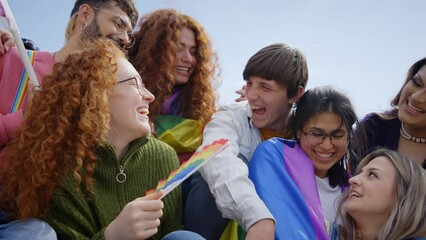Group laughing diverse people gathered sitting outdoor to demonstrate LGBT rights. Smiling happy friends enjoying together at pride party with rainbow flags and fans. Generation z and sexual freedom.