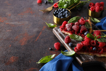 Pieces of berry pie on kitchen board, bowl with berries, basil, cutlery and towel on dark gray background, space for text