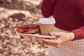 Woman walking in the park with cinnabon bun and coffee. Aesthetic autumn breakfast.