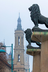 Fototapeta premium Statues of two lions guarding the entrance to the Stone Bridge in Zaragoza. Basilica of Nuestra Señora del Pilar in the background