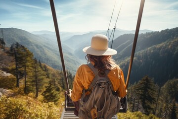 A young woman in a hat and with a backpack is walking on a suspension bridge in the mountains, Hiker woman riding a trolley zipline on the mountain, rear view, no visible faces, AI Generated