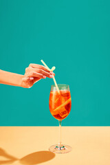 Female hand putting straw into Aperol spritz cocktail isolated over green background. Refreshment, aperitif. Concept of alcohol drinks, party, holidays, bar, mix. Poster. Copy space for ad - obrazy, fototapety, plakaty
