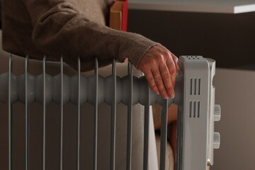 Woman in warm clothes is sitting next to the radiator. Woman is reading a book and warming her feet on an electric heater. It's cold at home. Energy crisis and high heating prices.