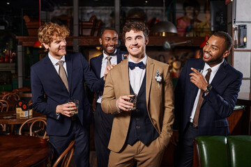 bachelor party, happy interracial best men looking at groom in suit standing with whiskey in bar