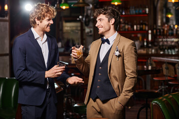 happy groom in suit with boutonniere and best man holding glasses of whiskey during bachelor party
