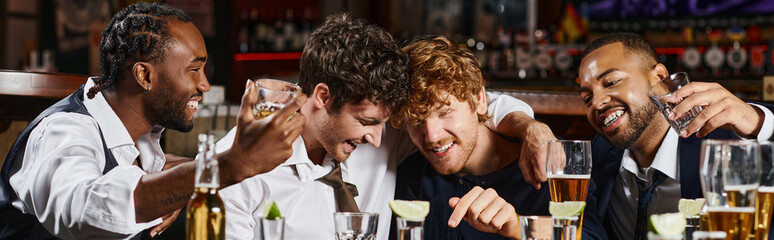 happy and drunk multiethnic friends hugging during bachelor party in bar, alcohol drinks banner