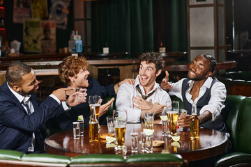 happy multiethnic men pointing at friend in bar, male friends during bachelor party in bar
