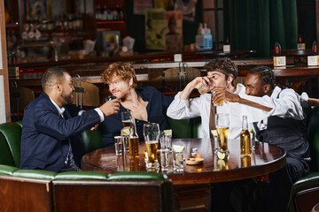 four happy multicultural colleagues drinking whiskey near tequila shots and beer on table in bar