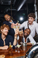 four happy multicultural friends toasting with glasses of whiskey near tequila shots and beer in bar