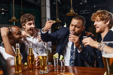 happy interracial men looking at african american friend drinking tequila shot and holding lime