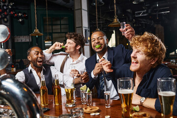 group of four happy and drunk multiethnic friends in formal wear drinking tequila in bar after work