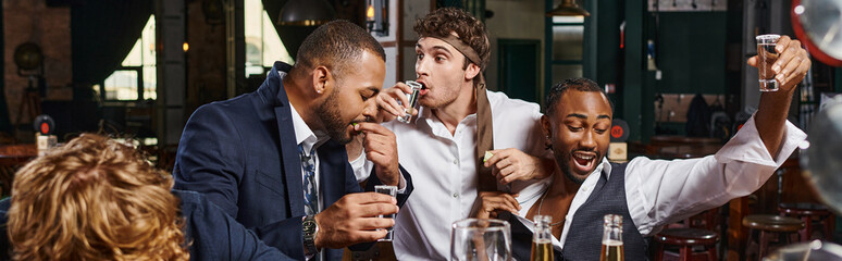 banner of funny and drunk multicultural men in formal wear drinking tequila in bar after work - obrazy, fototapety, plakaty