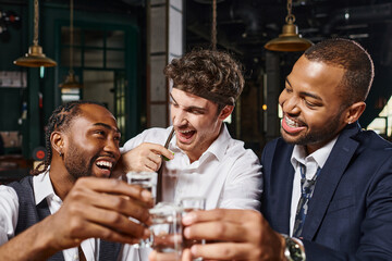 happy interracial friends toasting with glasses of tequila shots and laughing during bachelor party