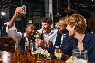 happy african american man raising shot of tequila near excited friends during bachelor party in bar