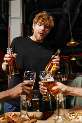 excited redhead man with curly hair holding bottles with beer near friends toasting glasses in bar