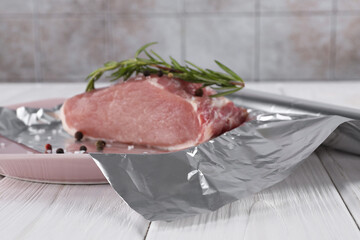 Aluminum foil with raw meat, rosemary and spices on white wooden table