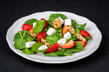 Fresh summer salad with strawberries, baby spinach, walnuts and feta cheese in a plate on a dark background. Concept for elegant presentation of healthy food.