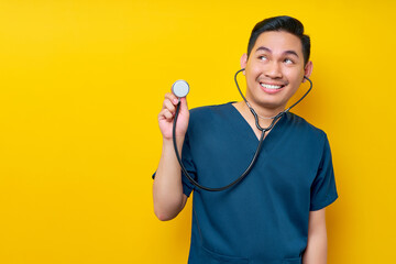 Professional young Asian man doctor or nurse wearing a blue uniform demonstrates using stethoscope isolated on yellow background. Healthcare medicine concept