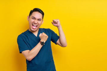 Excited professional young Asian male doctor or nurse wearing a blue uniform standing confidently and celebrating success isolated on yellow background. Healthcare medicine concept - obrazy, fototapety, plakaty