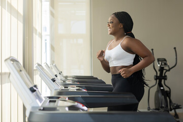 Side view of Cheerfully smiling plus size black woman in sportswear running on the treadmill in the...