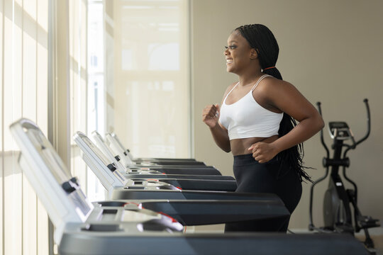 Side view of Cheerfully smiling plus size black woman in sportswear running on the treadmill in the modern gym. Morning begins with exercises